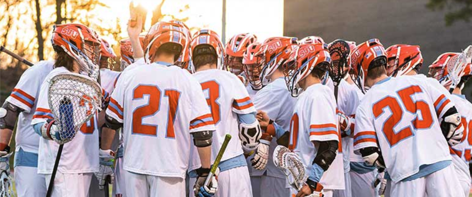 A lacrosse team dressed in white jerseys with red and blue accents huddles together on the field, with their sticks raised and helmets on. The sunset creates a glowing backdrop, giving the scene a warm and determined atmosphere.